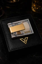 Vault Gold Money Clip by GoldPrivé holding folded US 100 dollar notes on a black leather box beside the emblem