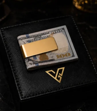 Vault Gold Money Clip by GoldPrivé holding folded US 100 dollar notes on a black leather box beside the emblem